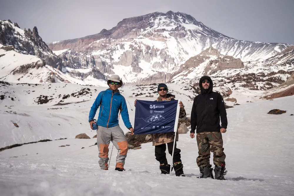 Fotografía de tres hombres mostrando una bandera con letras blancas en una zona nevada del volcán Tupungato en Chile.